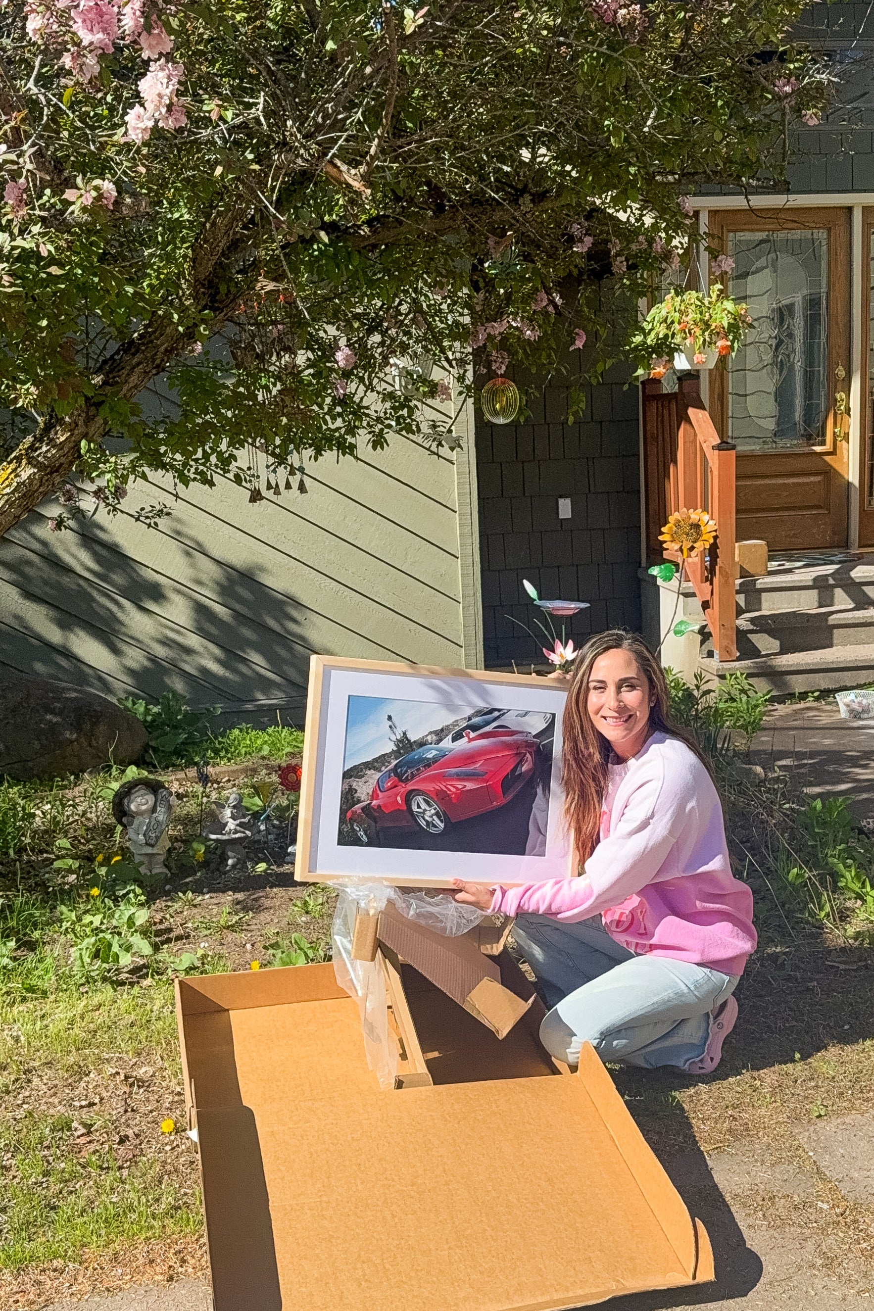 Woman holding a framed picture of a red car in front of a house with a tree and garden.